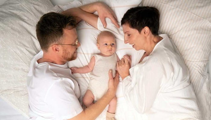 Father and mother with infant baby in bed white sheets 